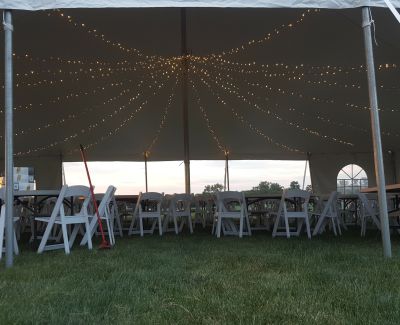 Large open tent with string lights and white folding chairs on a grassy lawn during evening setup.