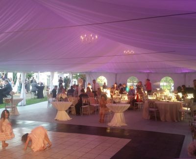 Outdoor wedding reception under a large white tent with chandeliers and purple lighting, guests seated at round tables.
