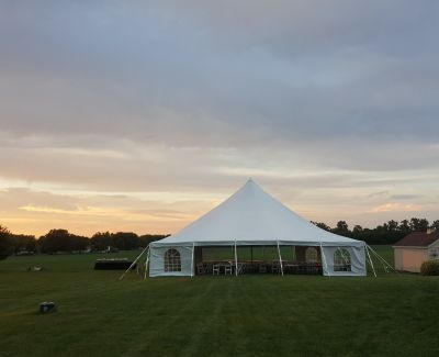 White high-peak wedding tent set on a green field at sunset with chairs and tables inside.