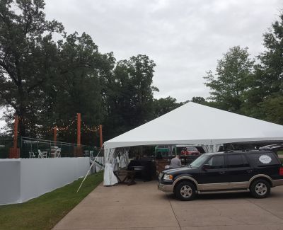 White canopy tent covering a driveway with parked cars beneath, surrounded by trees and fencing.