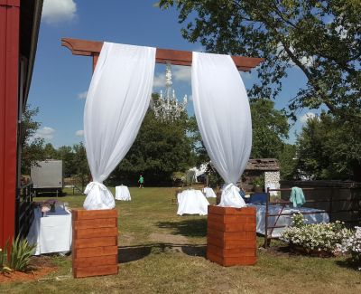 Outdoor wedding entrance with white draped archway near a barn, leading to cocktail tables and garden decor.
