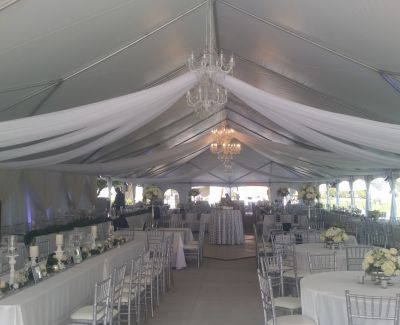 Elegant wedding setup inside a white tent with draped fabric ceiling, chandeliers, and white floral centerpieces.