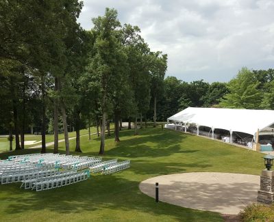 Outdoor wedding ceremony seating in a wooded area with a large event tent in the background.